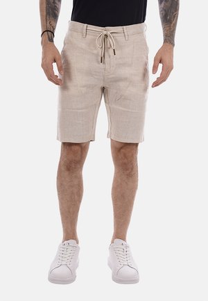Uomo con pantaloncini beige con coulisse, t-shirt nera, scarpe da ginnastica bianche e mostra tatuaggi su entrambe le avambraccia su sfondo semplice.