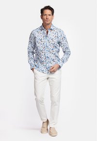 Chemise blanche à boutons avec un motif floral coloré ; manches longues, col classique. Associée à un pantalon blanc et des chaussures beiges.