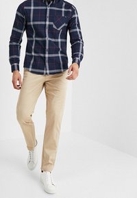 Camicia a quadri blu con linee bianche e rosse, taschino sul petto; abbinata a pantaloni chinos beige e scarpe da tennis bianche.