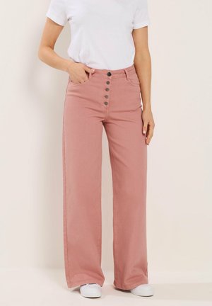 Kobieta ubrana w różowe spodnie z wysokim stanem i szerokimi nogawkami z pięcioma guzikami, białą koszulkę i białe trampki, stojąca na neutralnym tle.