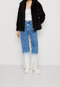 Henkilö, jolla on päällään musta pörröinen takki, valkoinen toppi, siniset ja valkoiset liukuväri leveät housut sekä valkoiset paksupohjaiset kengät.