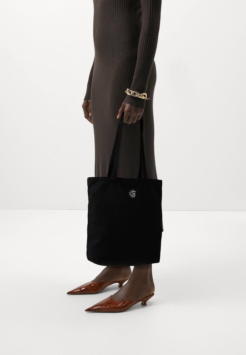 Sac en toile noir avec un emblème de soleil en blanc, doté de longues poignées. Le modèle porte une robe brune côtelée et des escarpins bruns à bout pointu.