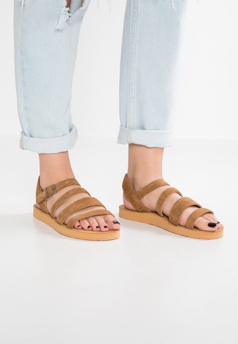Sandales en daim marron avec plusieurs brides horizontales, semelle beige clair, design à bout ouvert et fermeture à boucle sur le côté.
