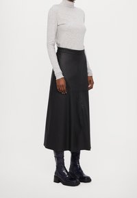 Langer schwarzer Ledermantel mit glatter Textur, kombiniert mit einem grauen Langarm-Rollkragenpullover. Das Modell trägt glänzende marineblaue Stiefel mit klobigem Absatz.