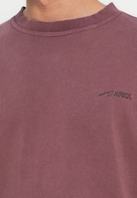 Εικόνα κοντινού πλάνου ενός ατόμου που φοράει ξεθωριασμένο μοβ T-shirt με στρογγυλή λαιμόκοψη και μικρό μαύρο λογότυπο «ARKK» στο αριστερό στήθος.