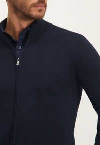 Marine blauwe rits-trui van gladde stof, met een hoge kraag, subtiel logo op de borst en een getailleerd ontwerp.