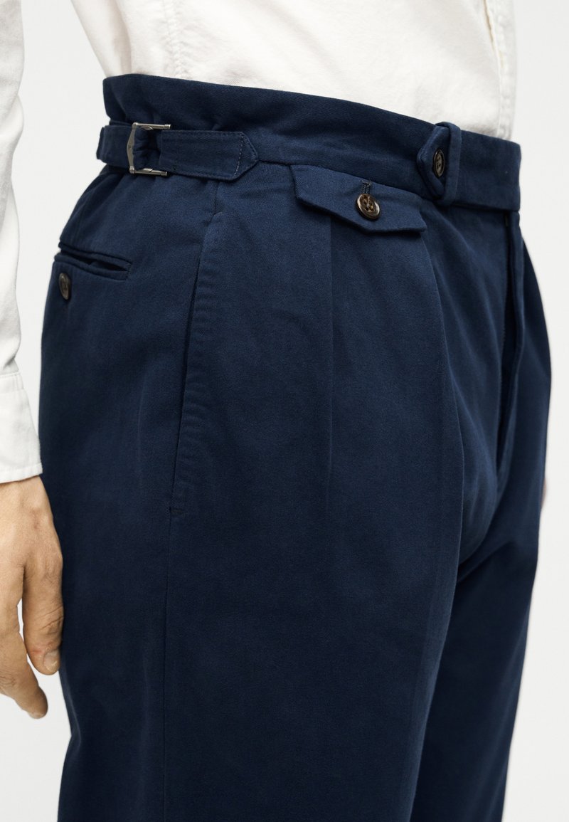 Pantalon bleu marine en tissu lisse, doté d'une ceinture avec une boucle argentée, d'un design plissé et de poches latérales.