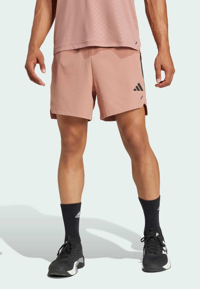 Shorts de sport roses avec des accents et un logo noirs, fabriqués à partir de matériaux légers. Portés avec des chaussettes noires et des baskets, mettant en avant un design sportif.