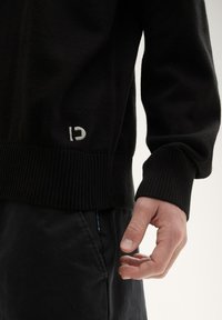 Svart stickad tröja med ribbad fåll och ärmslut, med en vit logotyp på sidan. Matchat med mörka byxor, med en närbild av en hand.