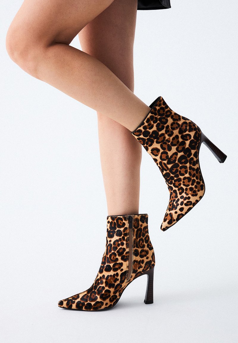 Mujer con botas de tacón alto de estampado de leopardo y cremallera lateral, un pie plano y el otro elevado contra un fondo blanco.