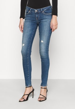Frau trägt enge blaue zerrissene Jeans, schwarzes langärmeliges Oberteil und schwarze Riemchen-High-Heel-Sandalen, steht vor einem schlichten Hintergrund.