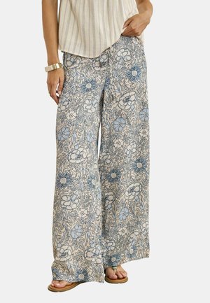 Persona que lleva pantalones de pierna ancha con un patrón floral azul y blanco, combinados con una camiseta beige a rayas, sandalias y una pulsera dorada.