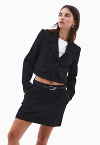 Blazer nero a righe con vestibilità corta, due tasche frontali e chiusura a un bottone, abbinato a una gonna coordinata e una cintura nera.