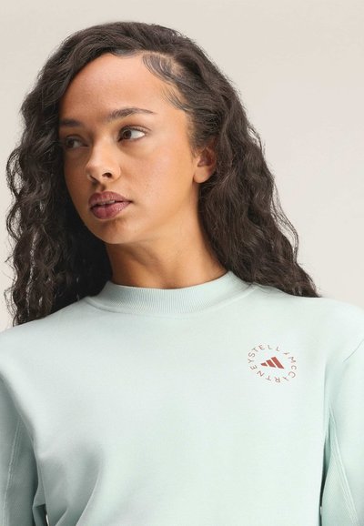 Kobieta o długich kręconych włosach, ubrana w jasnoniebieską bluzę z okrągłym czerwonym logo na piersi, patrząca w lewo na jednolitym tle.