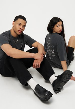 Two young adults sitting on the floor facing opposite directions wearing black casual outfits and chunky black shoes.