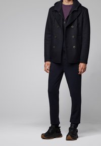 Marinblå dubbelknäppt sjömansrock med stora knappar, lager på lager över en mörk hoodie och en lila skjorta, kombinerat med mörka träningsbyxor och svarta sneakers.