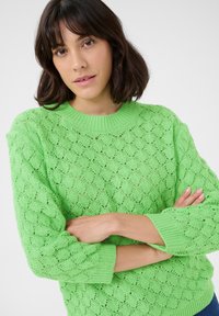 Pull en tricot vert vif, présentant un design en dentelle texturée avec un col rond et des poignets côtelés. Le motif évoque des vagues ou des coquillages.