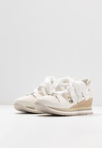 Vita kilklackar i texturerat läder, med elastiska remmar och kontrasterande beiga vävda sulor. Slät, minimalistisk design med låg profil.