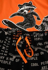 Pomarańczowa koszulka z grafiką bądź skateboardowym wiewiórkiem. Czarne spodenki z powtarzającym się wzorem tekstowym i pomarańczowym sznurkiem.