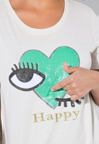 Camiseta blanca con un diseño de corazón de lentejuelas verdes, un gráfico de un ojo y la palabra "Feliz" en texto dorado. La textura es suave, con acentos brillantes.