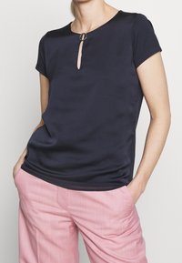 Marinblå kortärmad topp i slätt tyg, med en nyckelhålssnittsdetalj vid halsringningen med en cirkulär accent. Styler med rosa byxor.