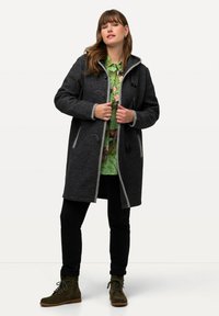 Cappotto in misto lana grigio scuro con cappuccio, bordi grigi, chiusure a toggle, tasche e una vestibilità comoda. Indossato sopra una camicia floreale verde e pantaloni neri.