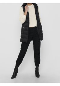 Person, der einen cremefarbenen Strickpullover, eine schwarze, gesteppte, ärmellose Daunenjacke, schwarze Cargo-Hosen und schwarze, hochhackige Stiefeletten trägt.