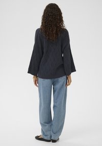 Marineblauer Strickpullover mit wellenförmigem Muster, weiten Ärmeln und lockerem Schnitt, kombiniert mit hellblauen, locker sitzenden Jeans und flachen Schuhen.