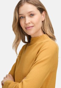 Mustársárga pulóver magas nyakkal és hosszú ujjakkal, puha kötött anyagból, egyszerű, varrás nélküli kialakítással, minták nélkül.