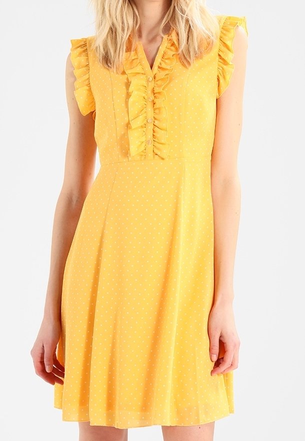 Femme portant une robe jaune sans manches à pois blancs, avec un col à volants et des boutons sur le devant.