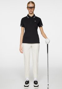 Svart polotröja med vit krage och detaljer, ihop med texturerade vita byxor. Modellen håller en golfklubba, iförd sportglasögon och handskar.