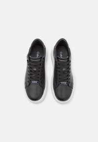 Pár černých nízkých tenisek s bílými podrážkami a černými tkaničkami, viděných shora na bílém pozadí.