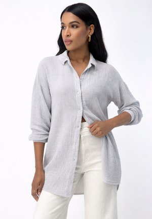 Kobieta z długimi ciemnymi włosami, ubrana w teksturowaną jasnoszarą koszulę na guziki i białe spodnie, patrzy na bok, jedną ręką poprawiając dół koszuli.