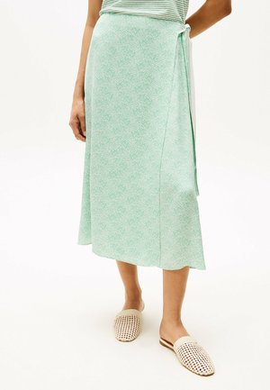 Groene wikkelrok met een gestructureerd patroon, van middellange lengte, met een zijstrik. Gecombineerd met beige geweven instapschoenen.