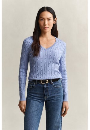 Frau mit langen dunklen Haaren, die einen hellblauen, kabelgestrickten V-Ausschnitt-Pullover und hochgeschnittene blaue Jeans mit schwarzem Gürtel trägt, vor neutralem Hintergrund stehend.