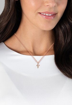 Et rose gold halskæde har en delikat kæde og et kors vedhæng dekoreret med små, klare ædelsten, båret over en hvid top.