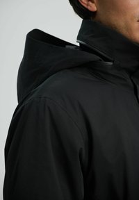 Chaqueta negra impermeable con un cuello alto. El diseño presenta una textura suave y un estilo minimalista, destacando la funcionalidad.