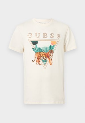 Tricou cu mânecă scurtă de culoare crem, cu un grafic cu tigru într-un triunghi, înconjurat de frunze verzi și textul "GUESS" deasupra.