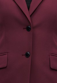 Σκούρο βυσσινί σακάκι με δύο μαύρα κουμπιά και τσέπες με καπάκι, φορεμένο πάνω από μαύρο τοπ.