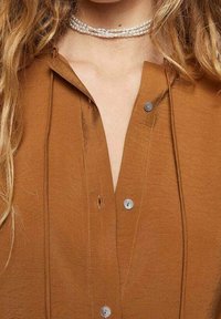 Bruine overhemd met knopen en een ontspannen kraag, voorzien van zilveren knopen en zichtbare stiksels. Choker ketting met parelaccenten.