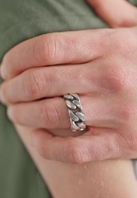 Sølv ring med et kjede-lignende design med glatte, avrundede seksjoner. Ringen er polert og står i kontrast til den teksturerte huden under.