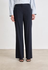 Calças largas de cor azul-marinho, feitas de um tecido suave, com uma cintura elástica e bolsos frontais, combinadas com sandálias pretas rasas.