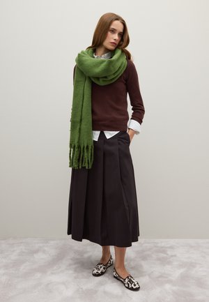 Maglione marrone, camicia bianca con colletto, lungo gonnellone nero e sciarpa verde oversize con frange. Scarpe con motivo in bianco e nero. Sfondo neutro.