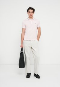 Ροζ ανοιχτό ροζ πόλο με σκούρες ρίγες, συνδυασμένο με ανοιχτό μπεζ παντελόνι. Το μοντέλο κρατά μια μαύρη τσάντα tote και φορά μαύρα παπούτσια.