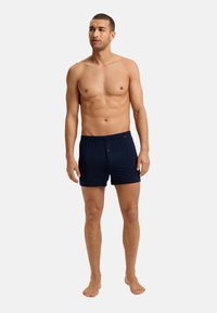 Boxer in cotone blu navy con vita elastica e dettaglio del bottone frontale. Texture liscia, vestibilità sartoriale e lunghezza della gamba corta.
