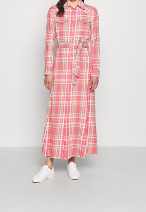 Femme portant une robe chemise à manches longues à carreaux roses et beiges avec une ceinture à la taille, associée à des baskets blanches, debout devant un fond uni.