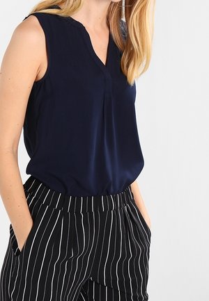 Vrouw draagt een mouwloze marineblauwe blouse, ingestopt in een zwarte broek met witte verticale strepen, handen in de zakken, tegen een effen achtergrond.
