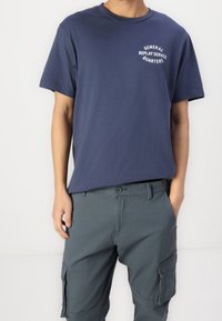 Granatowy bawełniany t-shirt z grafiką na lewej piersi z napisem „General Replay Service Quarters”, połączony z szarymi spodniami typu cargo z bocznymi kieszeniami.