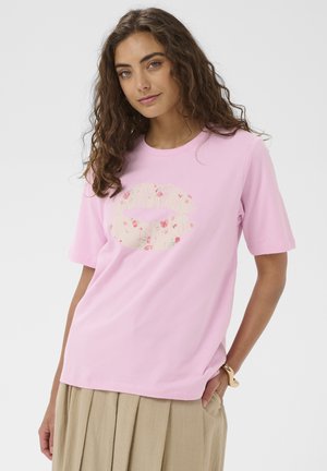 Fiatal nő göndör hajjal, világos rózsaszín, virágos szájnyomattal díszített pólóban és bézs pliszírozott szoknyában, az egyik kezét a zsebébe dugva áll.