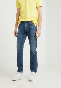 Ljusblå denimjeans med rak passform, kombinerat med en gul polo-tröja. Personen bär vita sneakers. Minimala detaljer i metall.